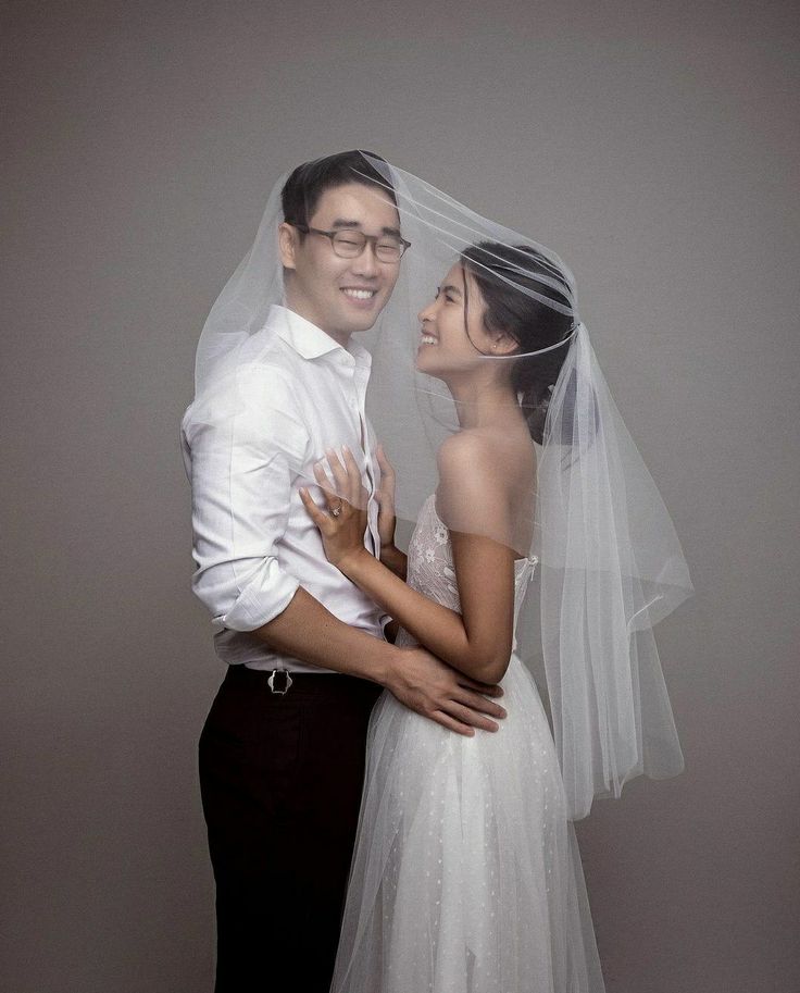 a bride and groom pose for a wedding photo in front of a gray background with their arms around each other