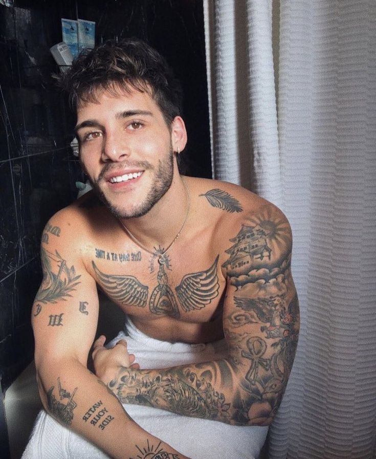 a man with tattoos on his chest sitting in front of a shower curtain and looking at the camera