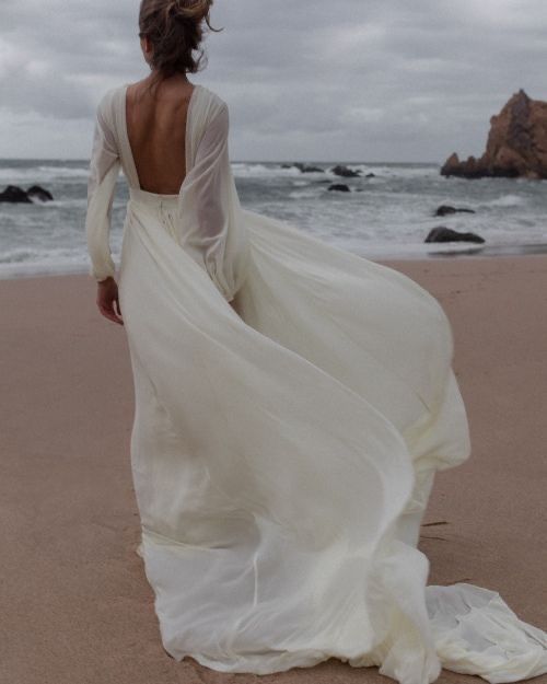 a woman in a long white dress walking on the beach with her back to the camera