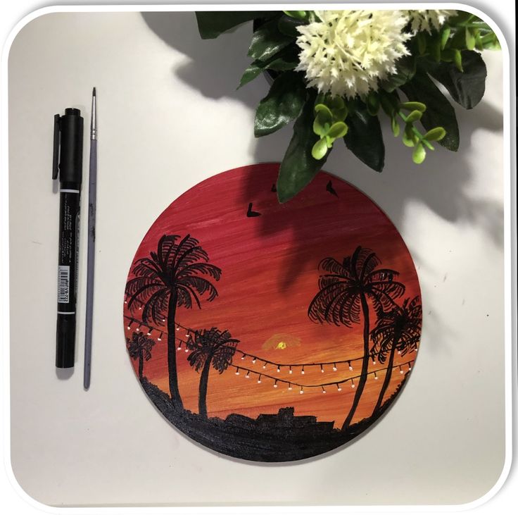 a wooden plate with palm trees painted on it next to a pen and flower pot