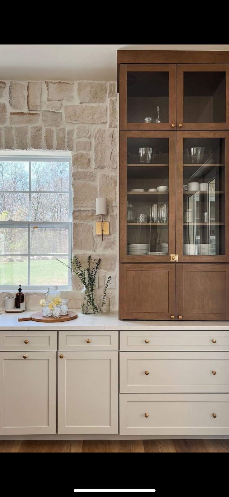 a kitchen with white cabinets and wood floors in front of a stone wall that has a window