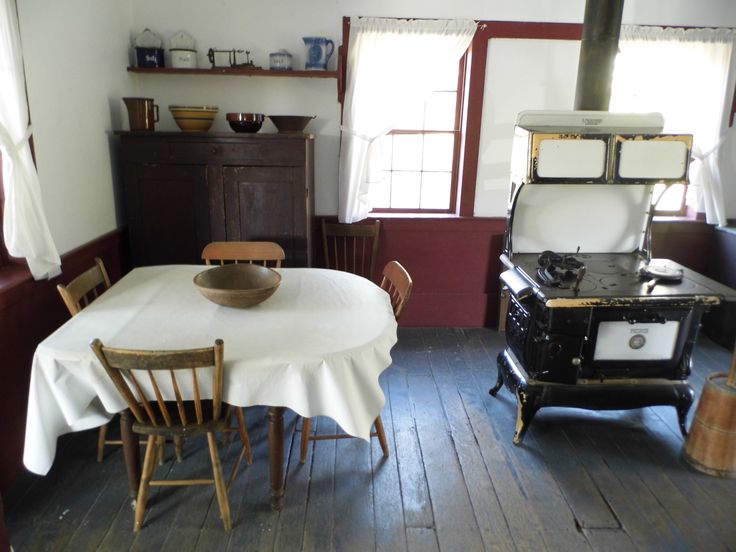 an old fashioned stove and table in a room with wooden floors, windows, and chairs