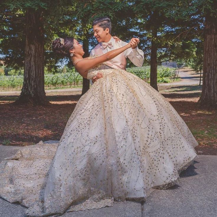 a man and woman dressed up in wedding gowns dancing on the street with trees behind them