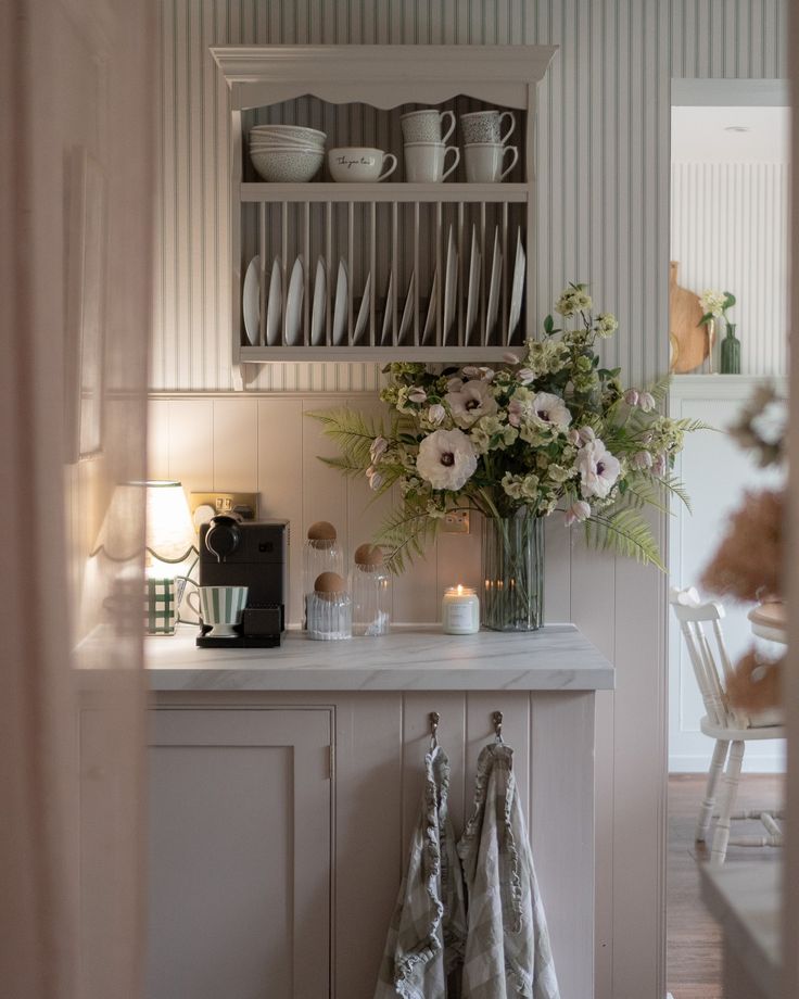 a kitchen with white cabinets and dishes on the shelves, flowers in a vase next to it