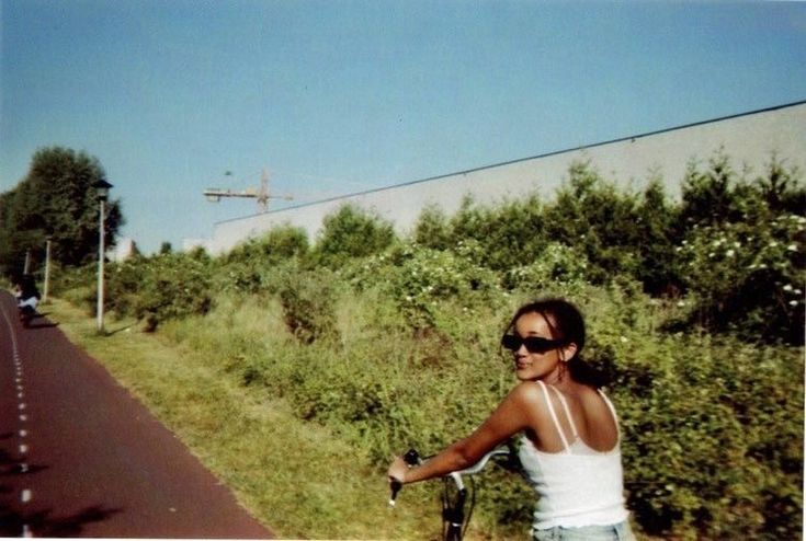 a woman riding a bike down a street next to a lush green field and tall grass