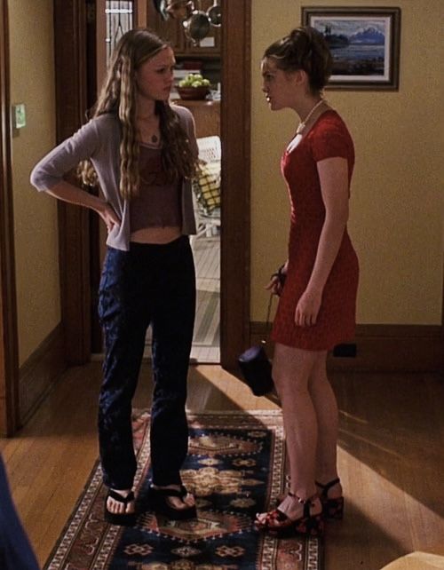 two young women standing in a living room talking to each other and one is holding her hand on her hip