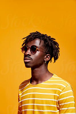 a man with dreadlocks standing in front of an orange wall wearing sunglasses and a striped shirt