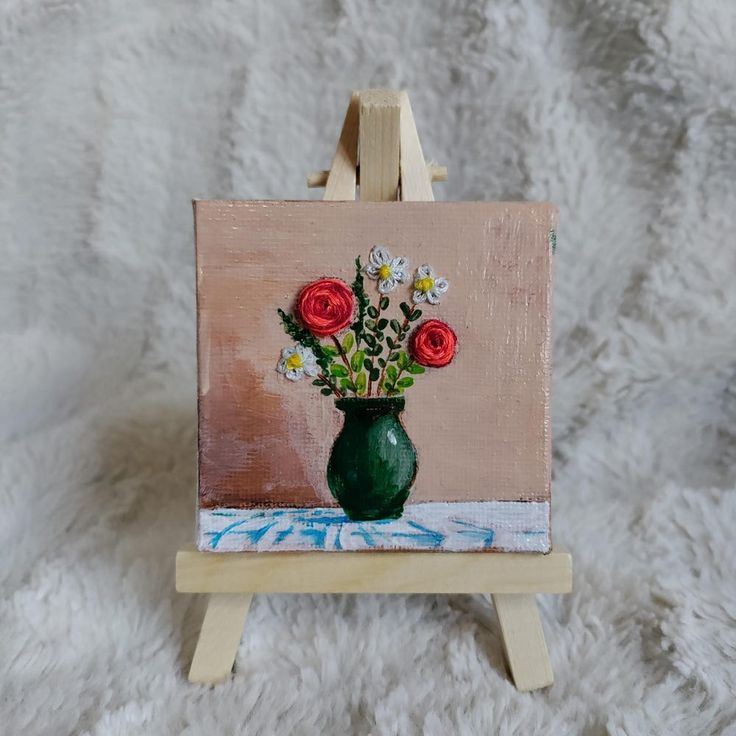 a small easel with a painting on it holding a green vase filled with red and white flowers