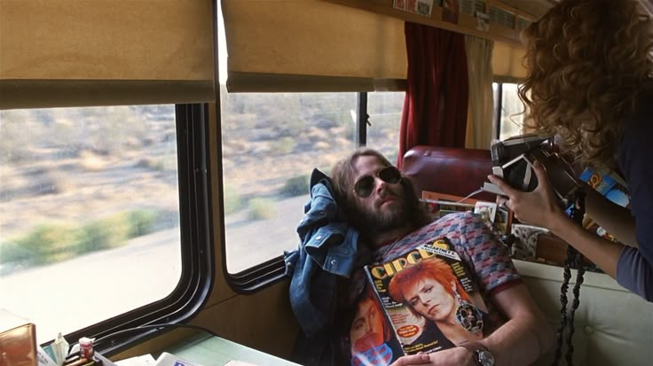 a man laying on top of a train next to a woman holding a camera and looking out the window