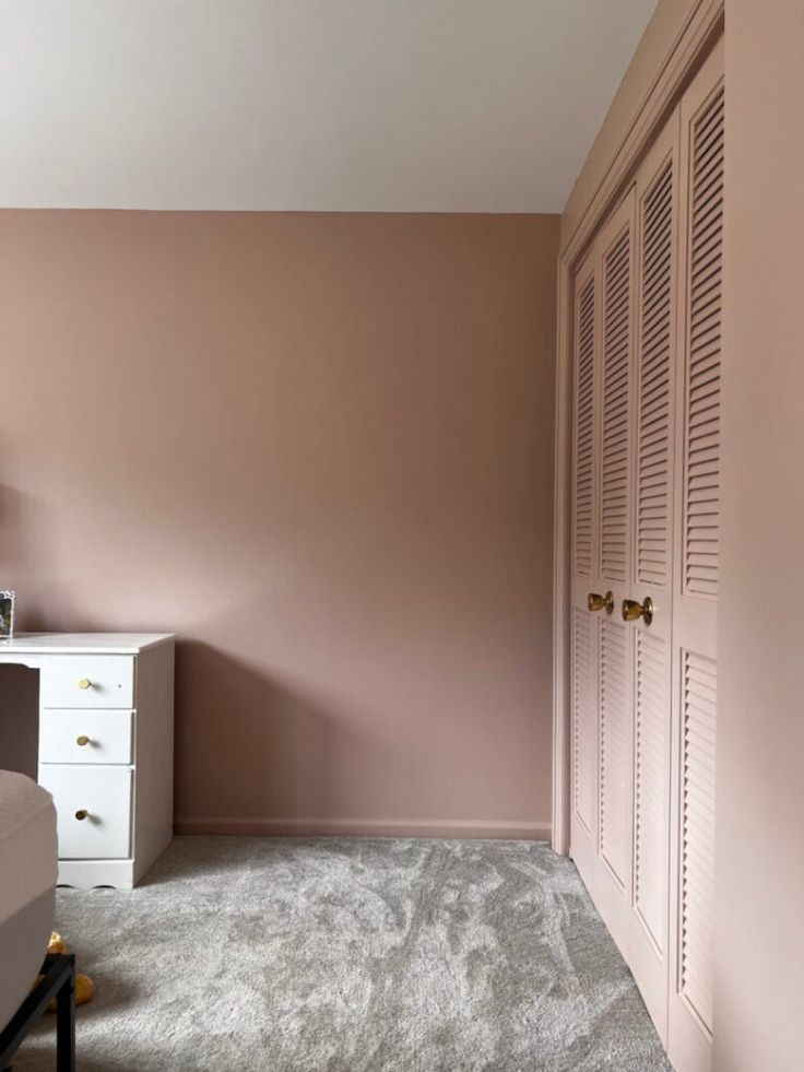 a bedroom with pink walls and carpeted flooring, white furniture and drawers in the corner