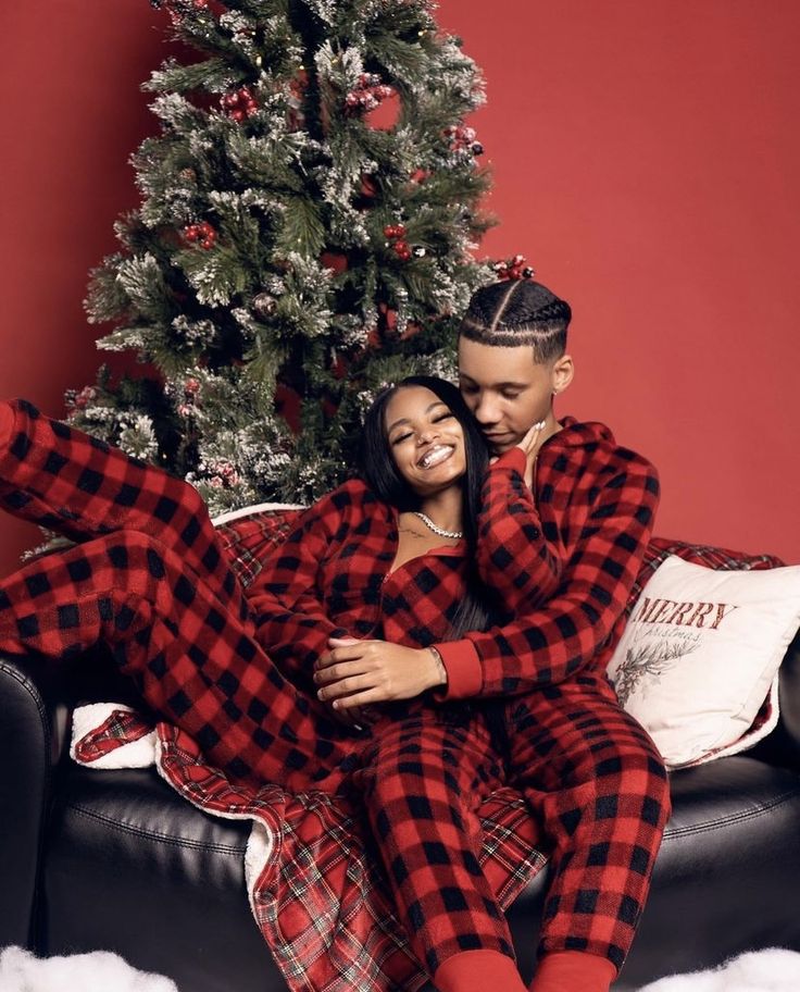 a man and woman in plaid pajamas sitting on a couch next to a christmas tree