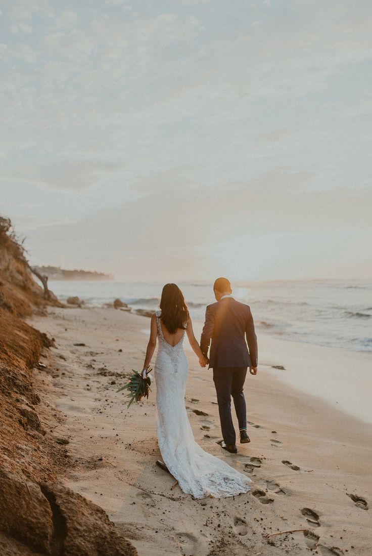 a bride and groom walking on the beach at sunset with their wedding dress blowing in the wind