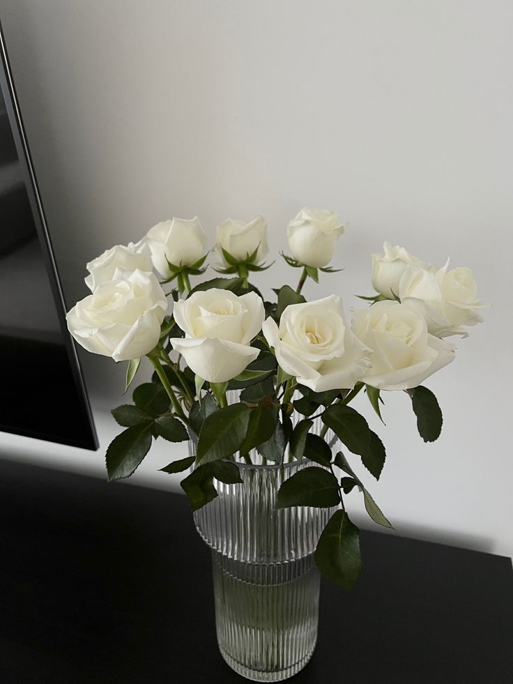 white roses are in a clear vase on a black countertop next to a mirror