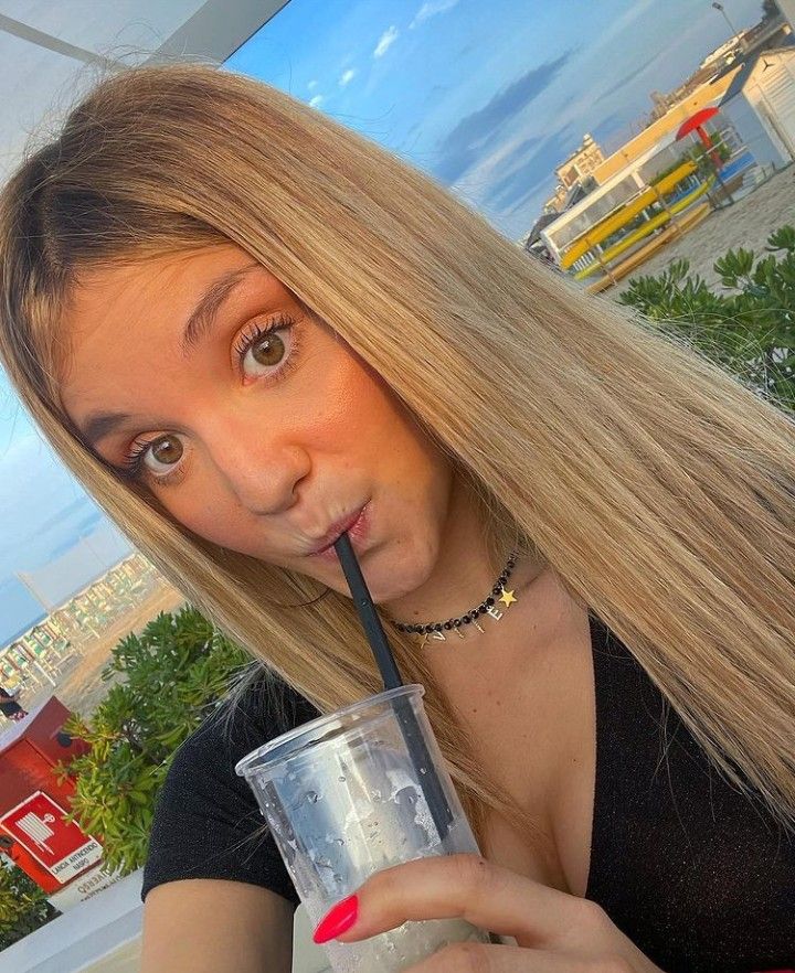 a woman with long blonde hair drinking from a straw in front of her face and looking at the camera