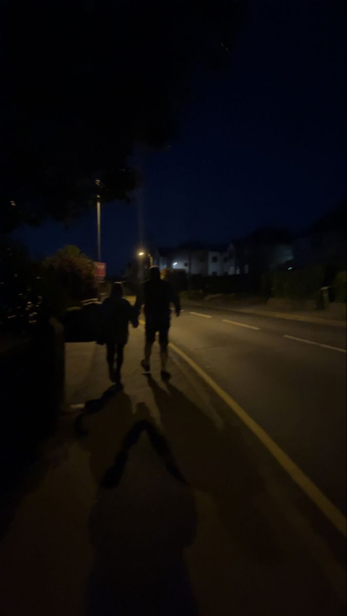 two people walking down the street at night with their shadows on the road and buildings in the background