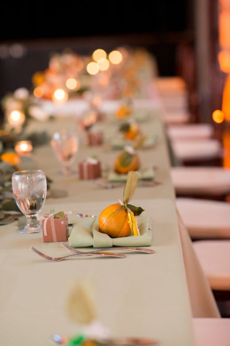 a long table is set with place settings and oranges on the plates, along with candles