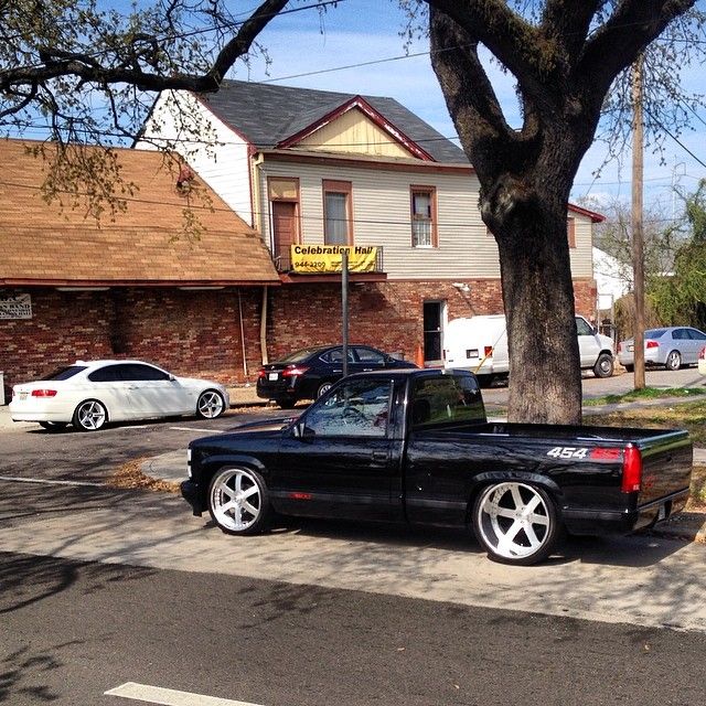 a black truck parked on the side of a road next to a tree and buildings