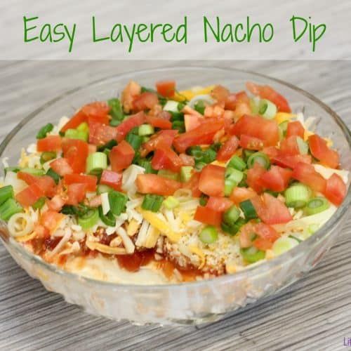 an easy layered nacho dip with tomatoes, onions and celery in a glass bowl