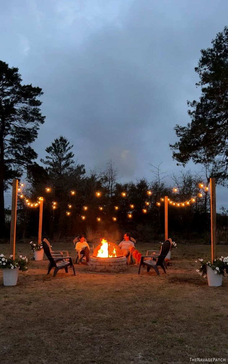 people sitting around a fire pit in the middle of a field with lights strung over it