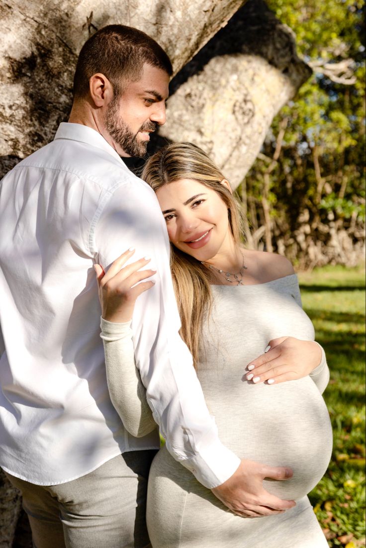 a pregnant couple standing next to each other under a tree