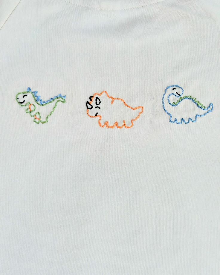 three embroidered dinosaurs on a white shirt with orange, blue, and green trims