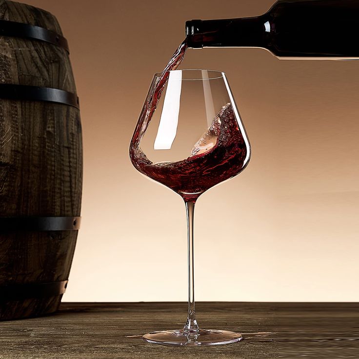 a wine glass filled with red wine being poured into it by a wooden barrel on the table