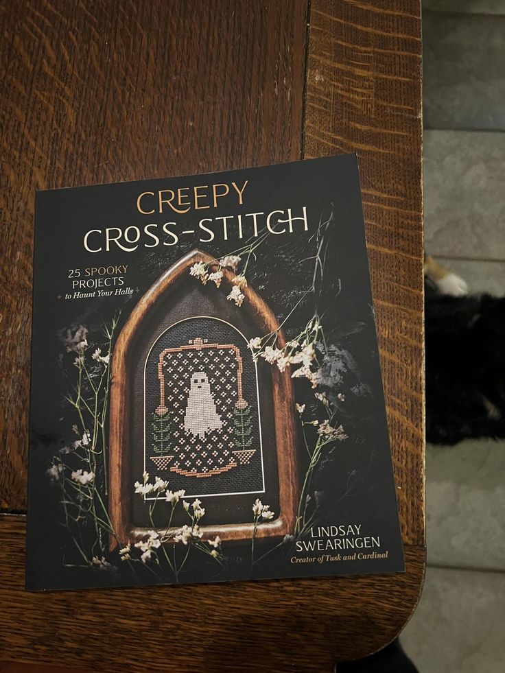 the book creepy cross - stitch is sitting on top of a wooden table next to a black cat