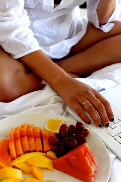 a woman in white shirt sitting on bed next to plate of fruit and laptop computer