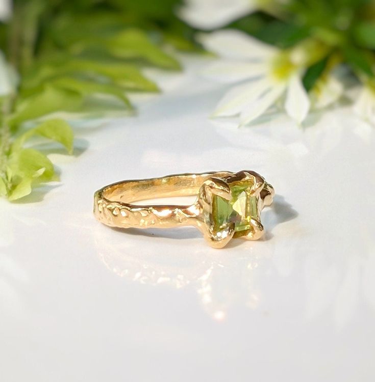 Show your love with this exquisite Peridot ring. Handcrafted in wax and cast in Solid 14k Gold, this stunning green crystal ring features an organic textured band, ensuring its uniqueness. Adorned with a mesmerizing princess-cut Peridot, also the August birthstone, it embodies more than an engagement ring - it symbolizes timeless love and individualityT H E ∙ S M A L L ∙ D E T A I L S• Princess-cut Peridot (5 x 5mm)• Solid 14k Gold textured bandM A T E R I A L ∙ O P T I O N S• Sterling Silver• S Peridot And Diamond Engagement Rings, Unique Yellow Gold Ring With Birthstone, Unique Yellow Gold Birthstone Ring, Elegant Peridot Emerald Promise Ring, Yellow Gold Peridot Jewelry With Emerald Cut, Elegant Green Citrine Jewelry, Tsavorite Birthstone Rings For Wedding, Wedding Jewelry In Yellow Gold With Tsavorite, Emerald Cut Peridot Gemstone Jewelry