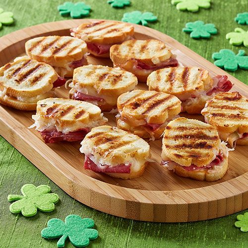 small sandwiches are arranged on a wooden platter with shamrocks in the foreground