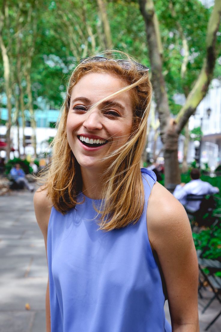 a woman with long hair smiling and wearing a blue top on the street in front of trees