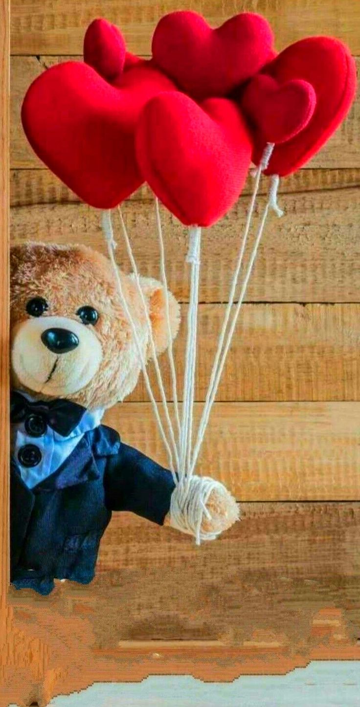 a teddy bear holding two red heart shaped balloons in its mouth while standing on a wooden wall