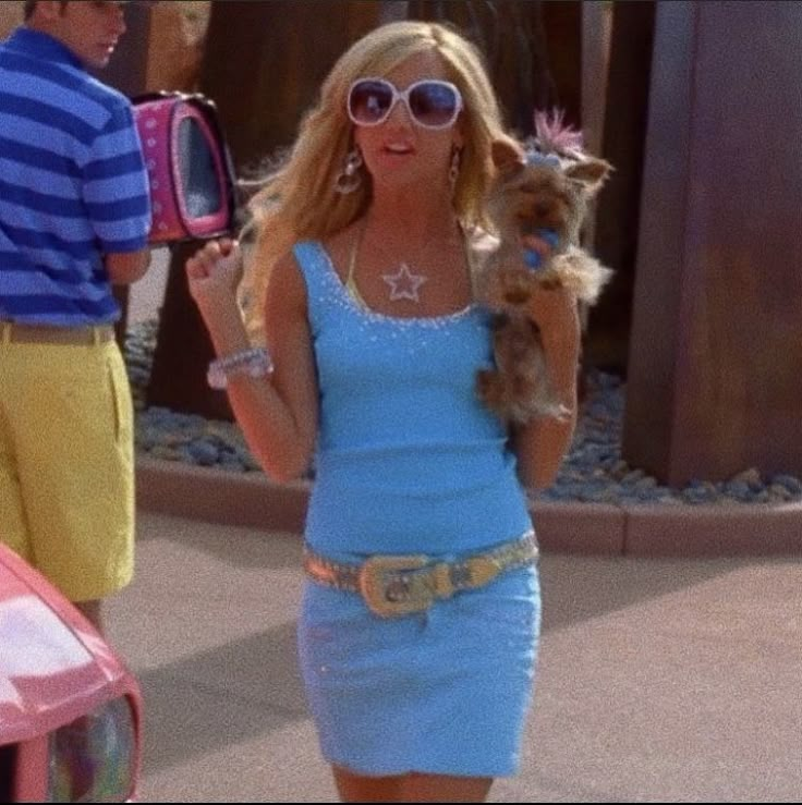 a woman in a blue dress holding a small dog on her hip while standing next to a pink car