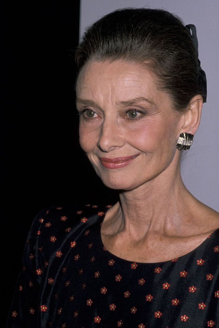 an older woman with big earrings on her head smiling at the camera while standing in front of a white wall