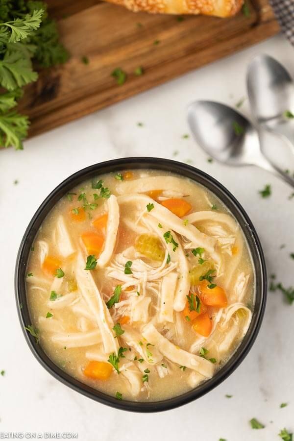 a bowl of chicken noodle soup with parsley on the side and bread in the background