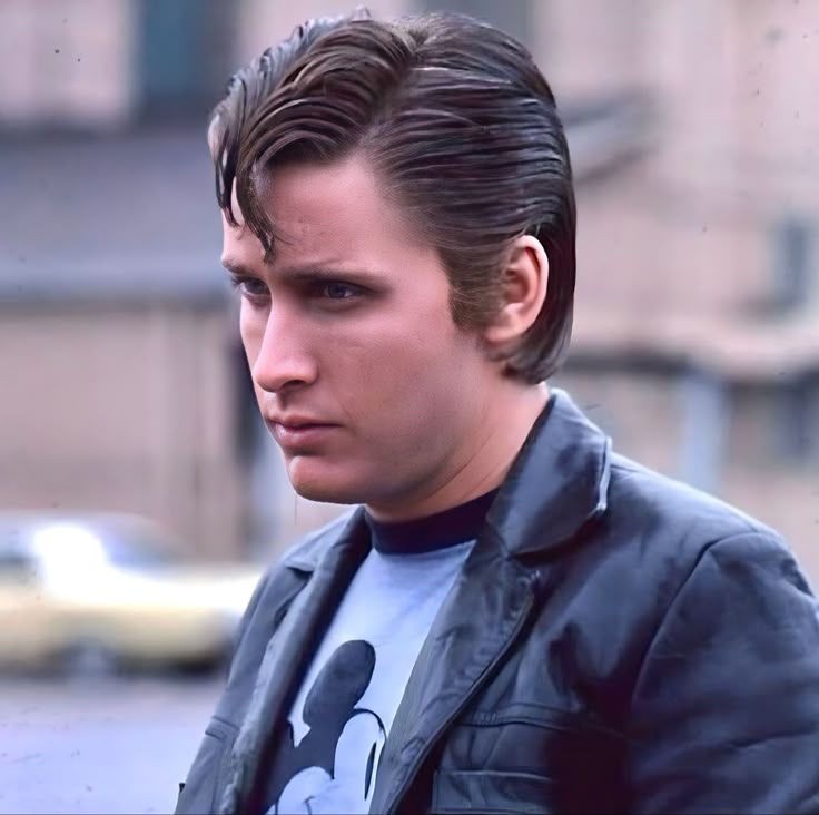 a young man wearing a mickey mouse t - shirt and leather jacket looking at the camera