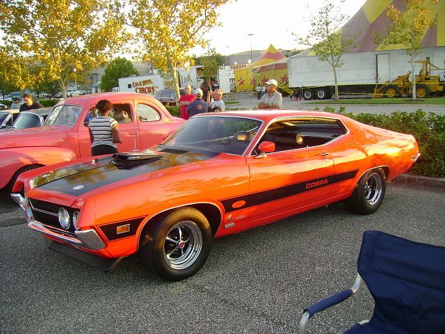 an orange muscle car parked in a parking lot next to other old cars and people