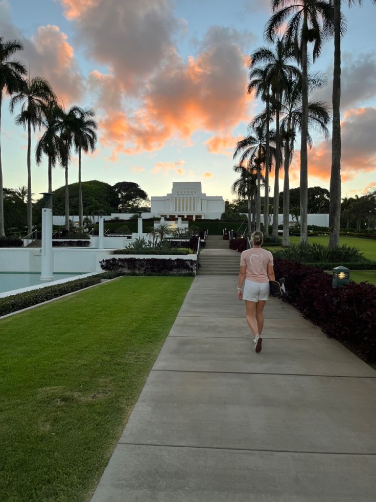 a woman walking down a sidewalk in front of palm trees and a white building at sunset