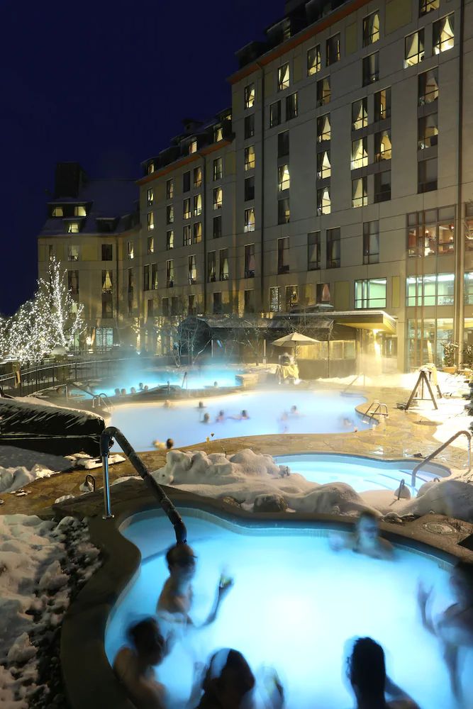 people are in an outdoor hot tub at night with snow on the ground and buildings behind them