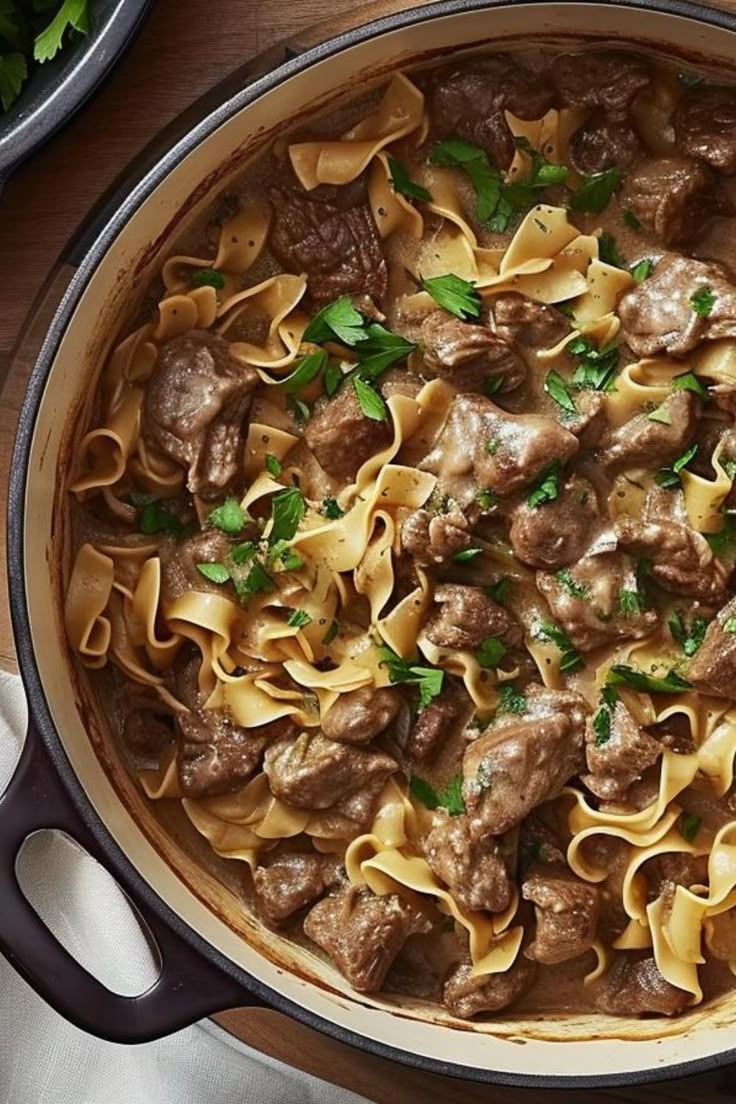 a pot filled with beef and noodles on top of a wooden table next to utensils