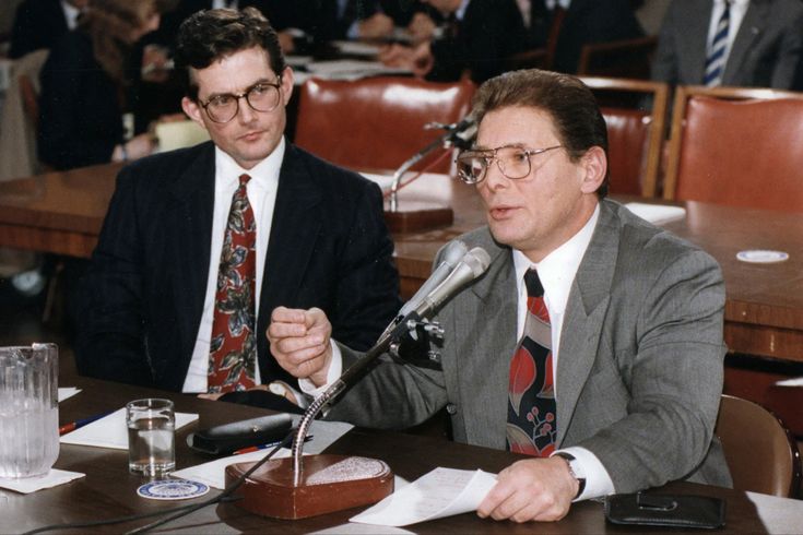 two men sitting at a table with microphones in front of them and papers on the table