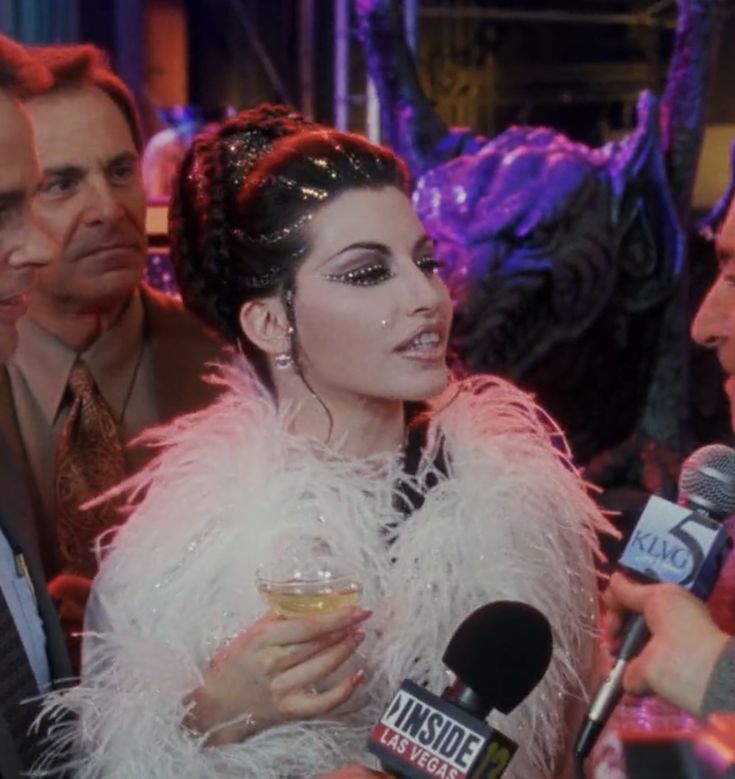 a woman in a white feather coat holding a microphone and talking to the camera while two men stand behind her