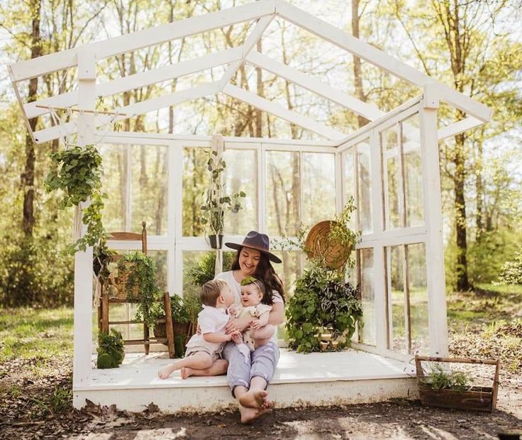 a woman holding a baby sitting in front of a white greenhouse with potted plants