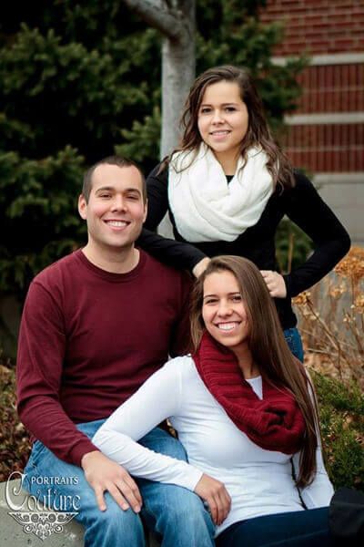 three people sitting on a bench in front of some trees and bushes, smiling for the camera