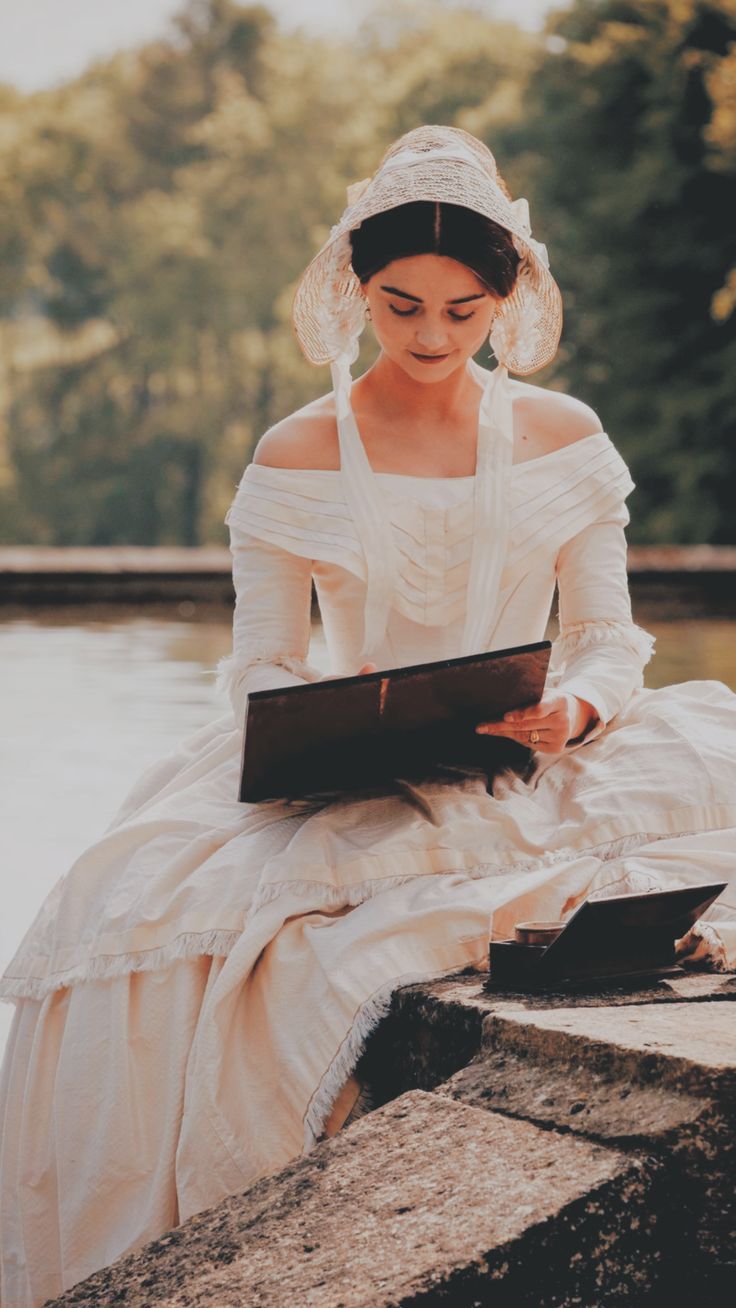 a woman dressed in white is sitting on the edge of a lake and reading a book