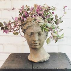 a statue head with purple flowers in it on top of a black box next to a brick wall