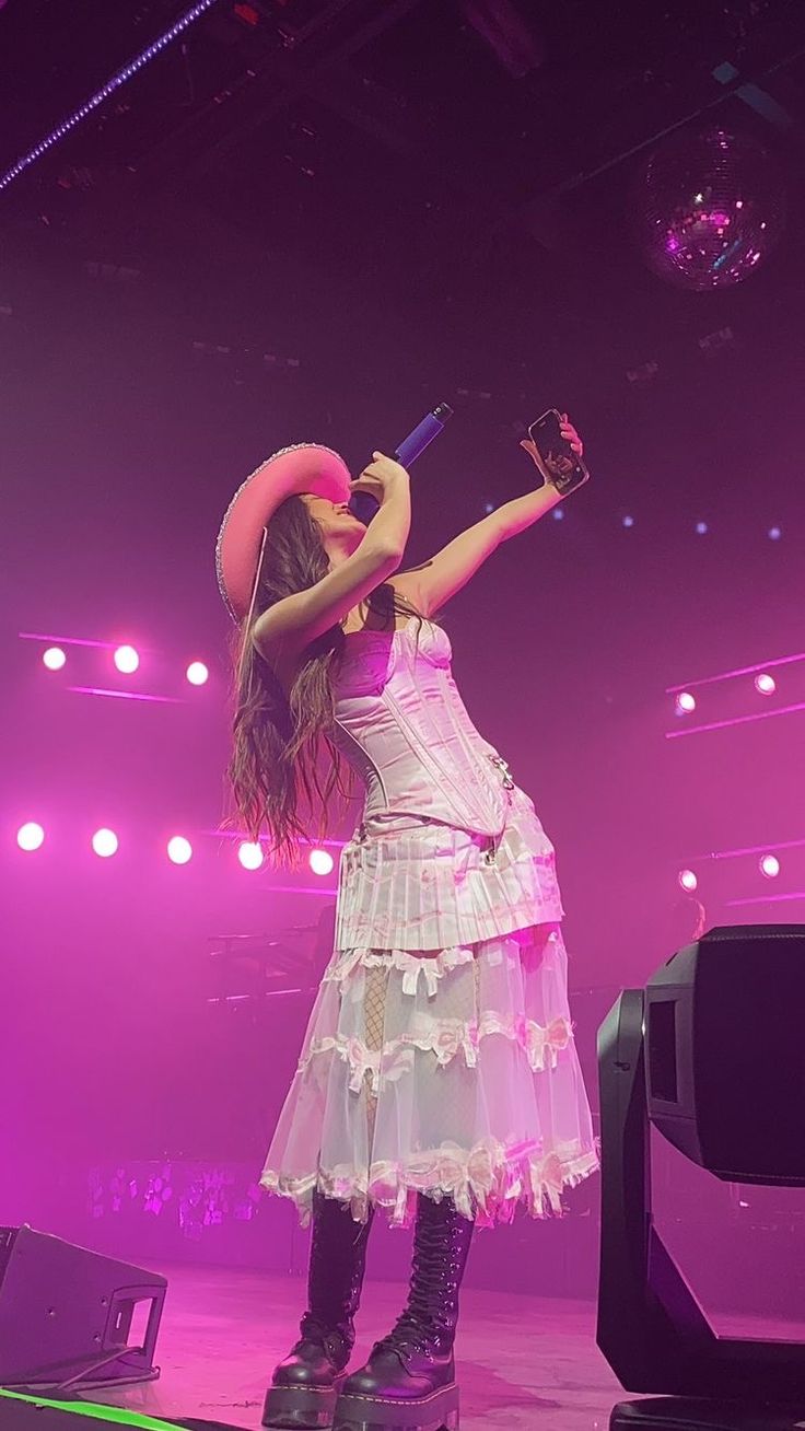 a woman in a white dress and hat on stage holding a pink ring around her neck