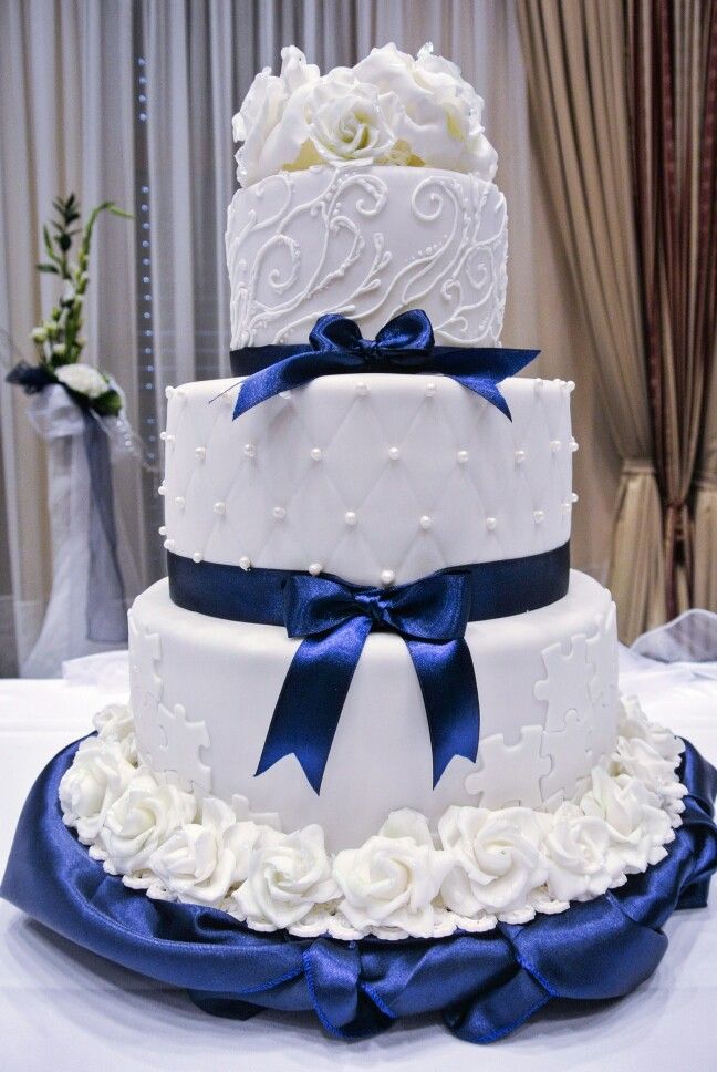 three tiered wedding cake with blue ribbon and white roses on the bottom, sitting on a table