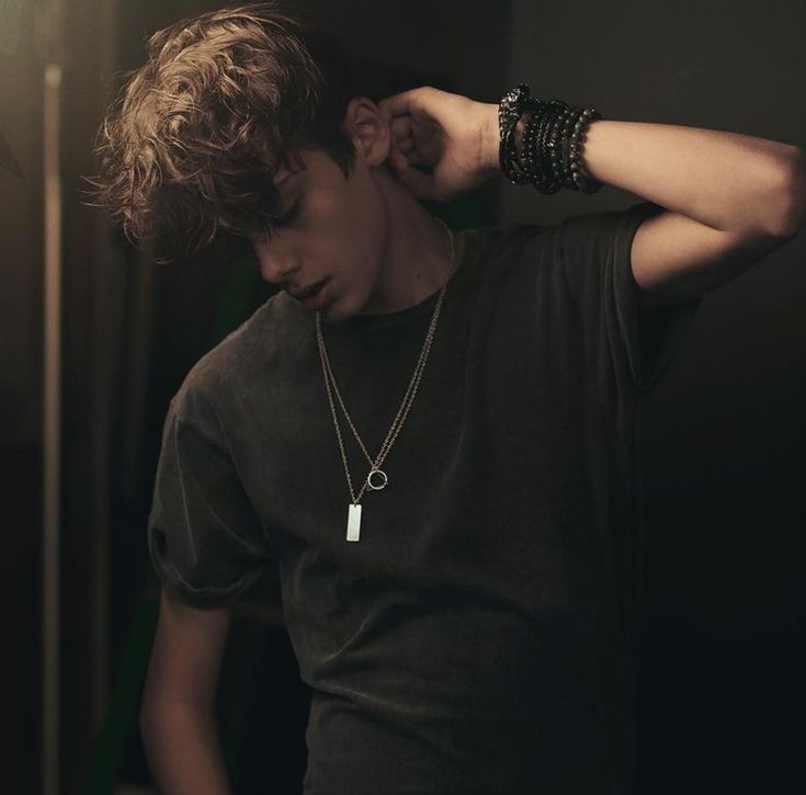 a young man wearing a black t - shirt and bracelets is leaning against a wall