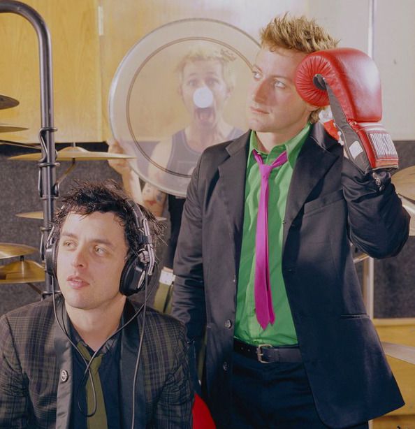 two men with headphones and microphones in front of a recording studio backdrop, one holding up a pair of red ear muffs